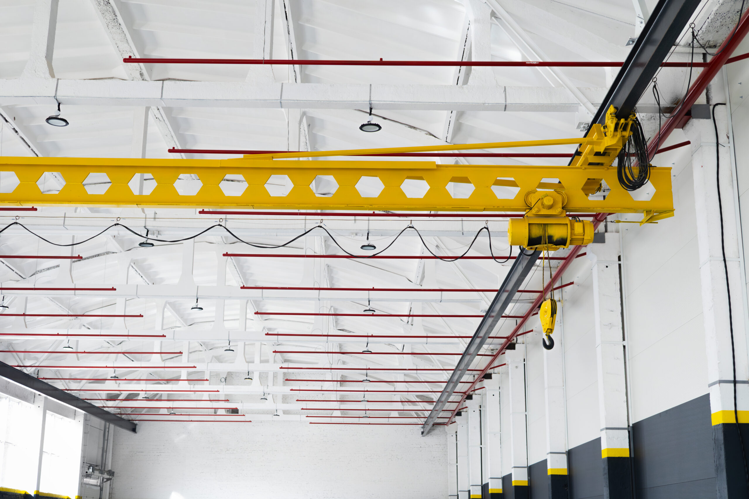 Yellow overhead crane with hook in a bright industrial hall. Steel beams and clean factory interior with visible red fire sprinkler system, heavy lifting equipment for manufacturing and logistics.