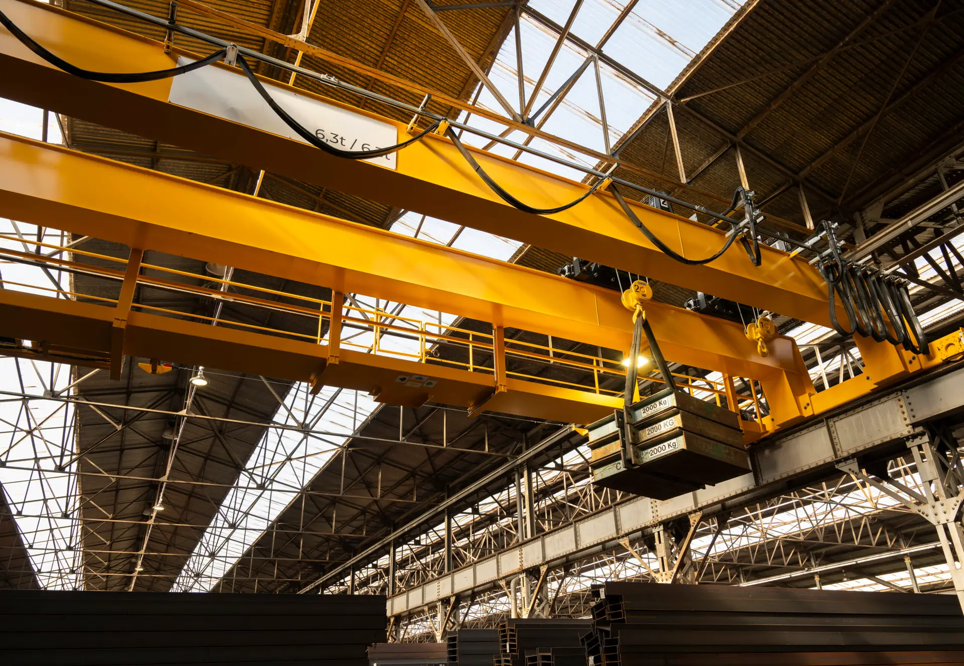 Heavy duty overhead crane lifting a stack of metal beams along its yellow track inside a large industrial manufacturing facility, with natural light filtering through the roof.